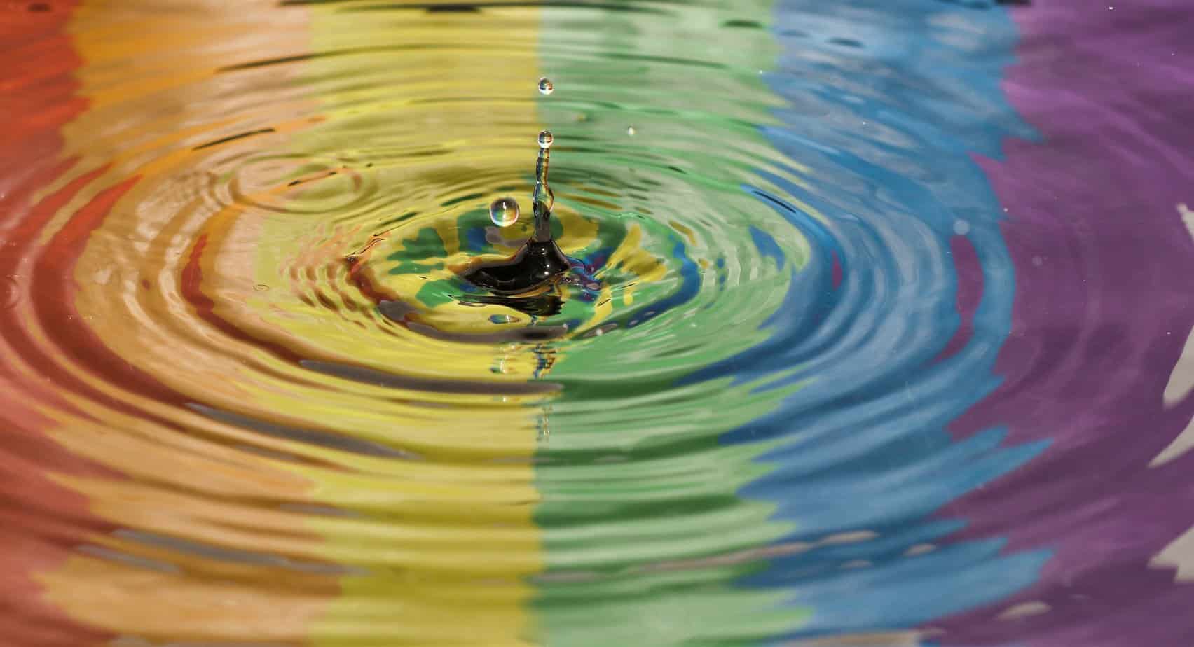 A puddle reflects a rainbow pride flag as a drop of water drips into it, creating ripples.