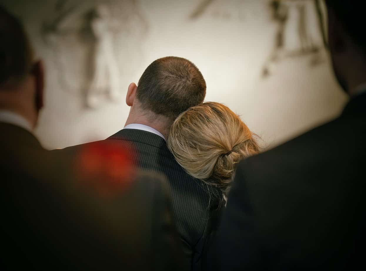 A woman rests her blonde head on the shoulder of a man in a dark suit, grieving