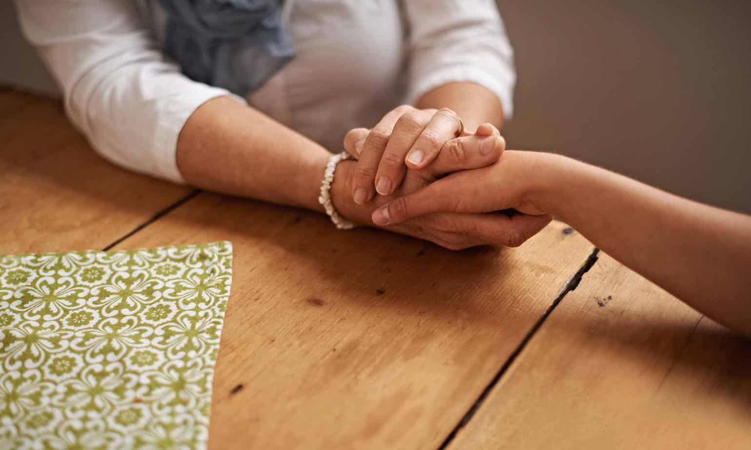 An older woman wearing a white shirt and bracelet holding a hand of a younger alcoholic on a wooden table at Mountainside Treatment Center