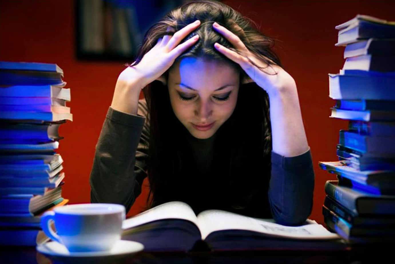 stressed woman with hands in her hair looking at book reading on desk with books piled by her with cup of coffee