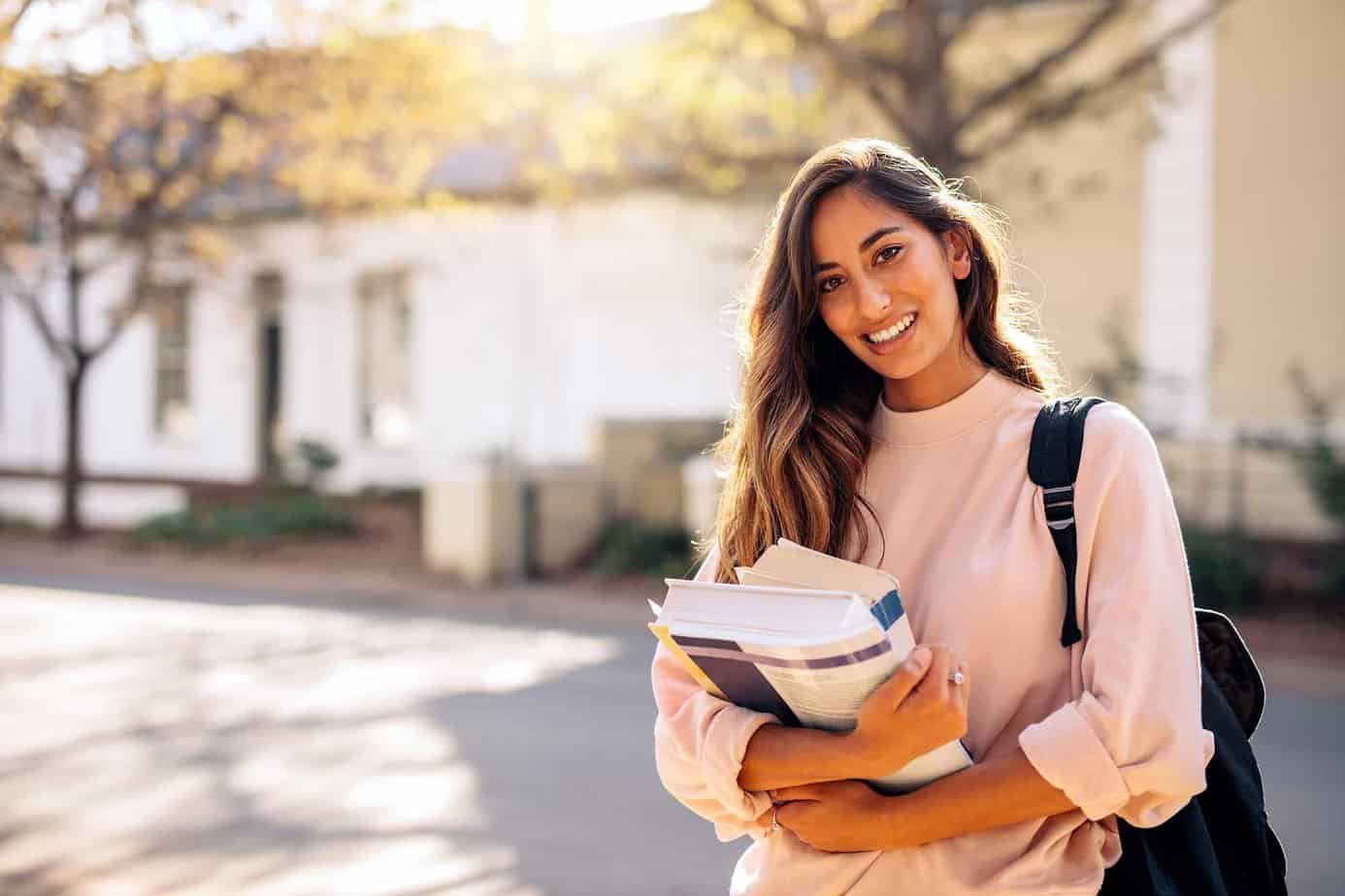 Female college student with backpack smiling while holding books at college campus
