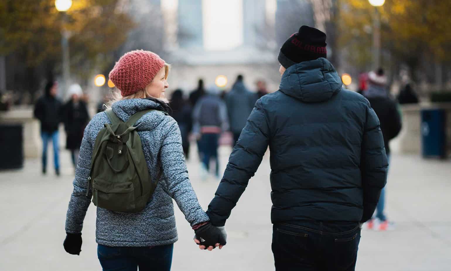 man and smiling woman in blue jackets and hats holding hands walking on a cold day