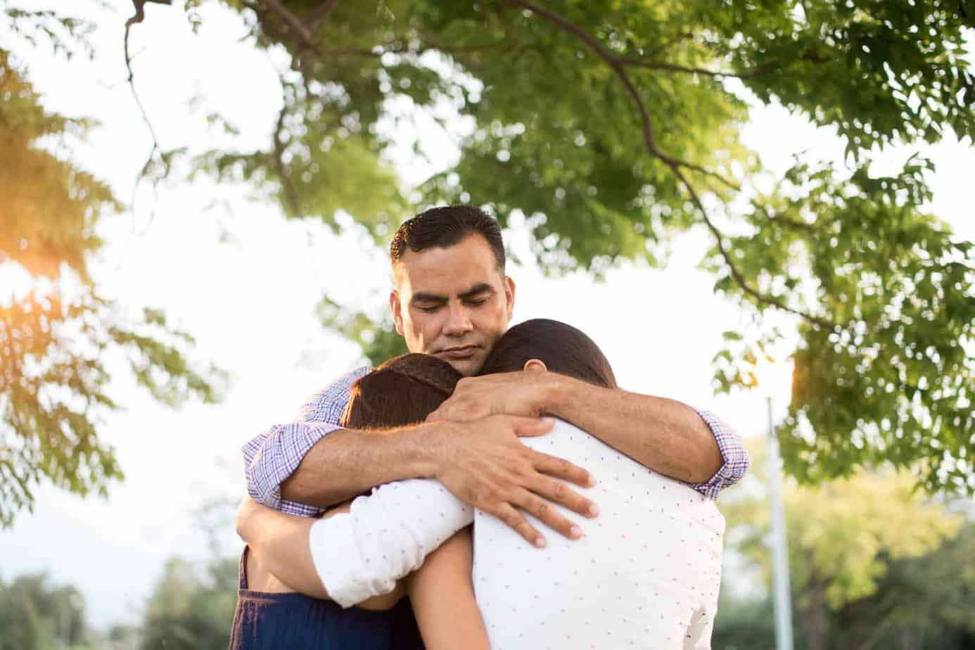sad man hugging two females outside under a tree
