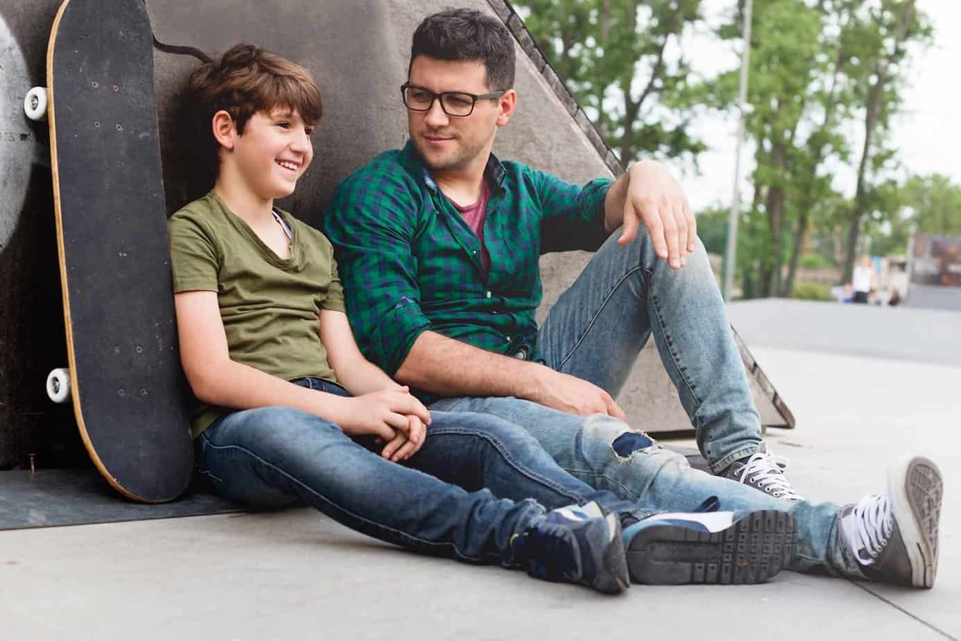 Man and boy sitting next to skateboard in park smiling