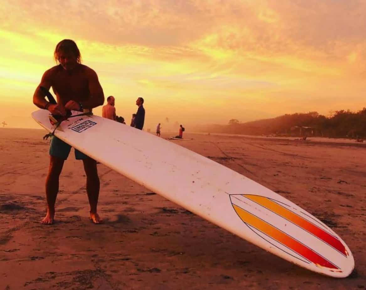 surfer prepping surfboard on beach sand during sunset