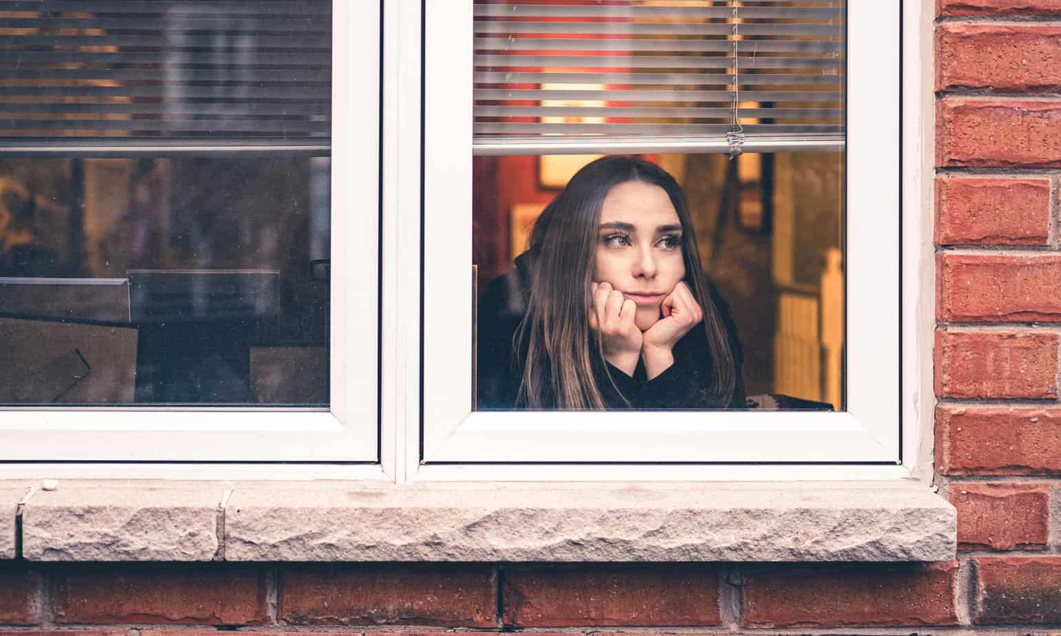 woman looking sad through a window with hands on face