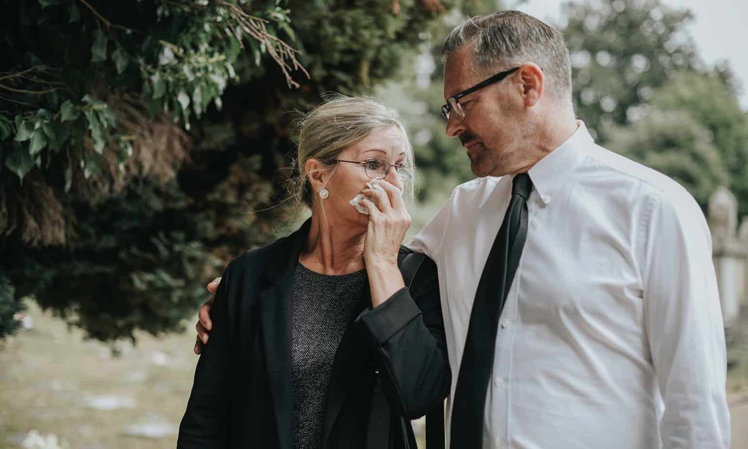 older woman in black covering face with tissue while older man in shirt and tie has arm around her