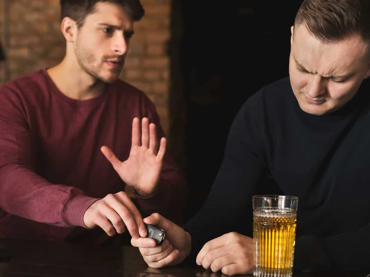 man in red shirt taking car keys away from sad man drunk with beer in front of him in a bar
