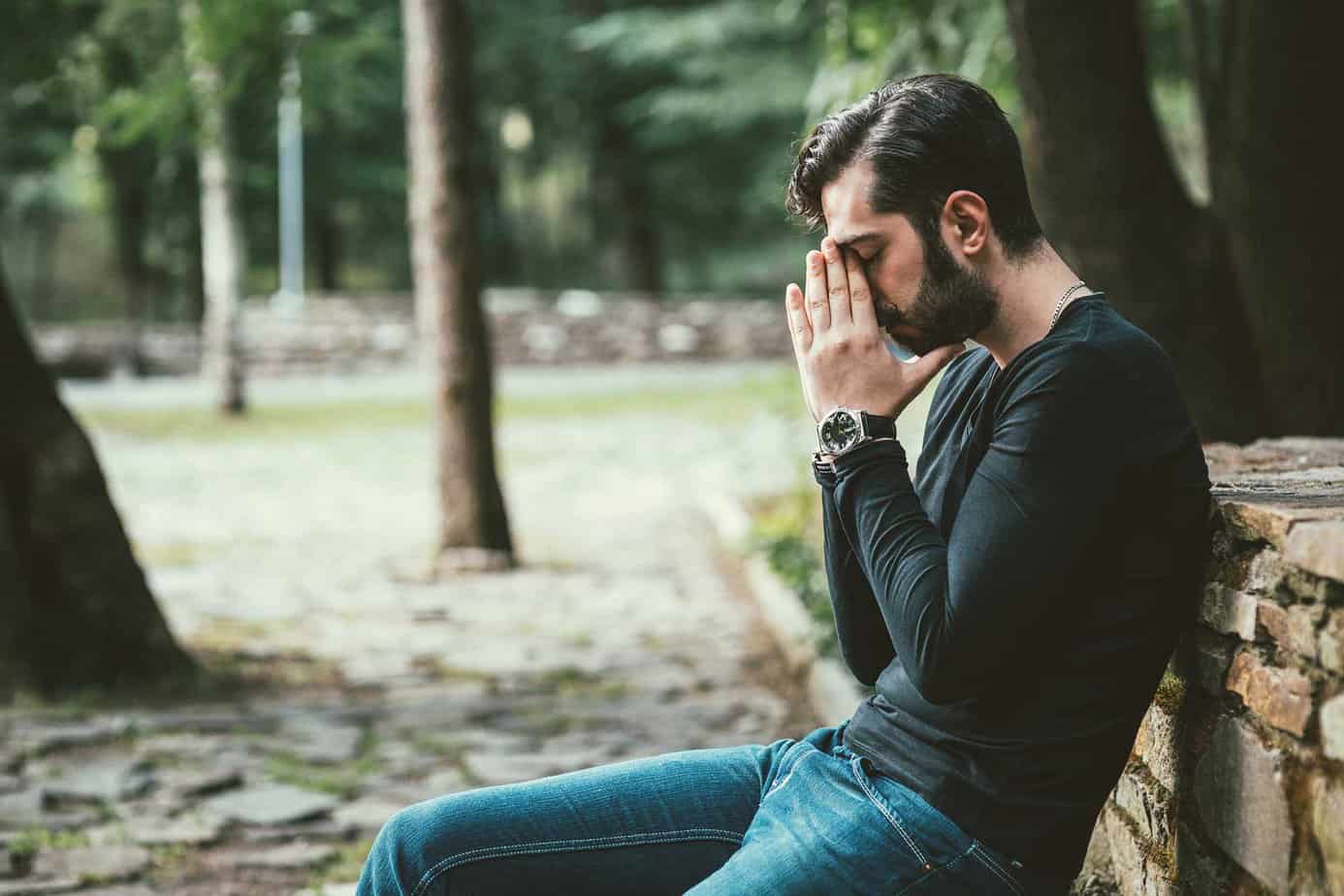 Man in long black shirt and jeans sitting on park bench outside near trees with hands on head in prayer pose