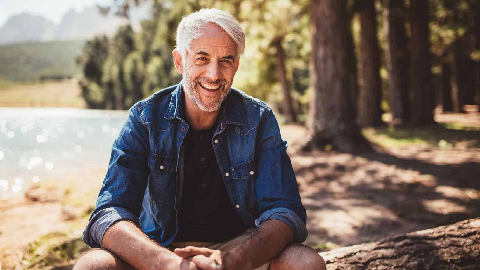 Older white haired man with denim shirt and shorts smiling sitting on log by lake and trees