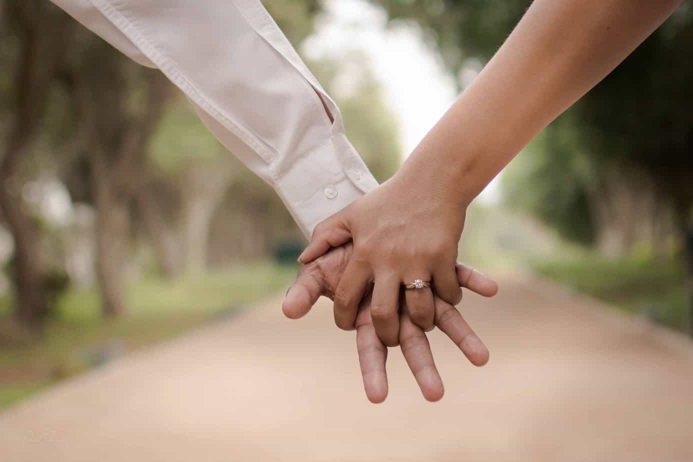 Close up of hands with man in button up white shirt and woman wearing engagement ring walking on path outside