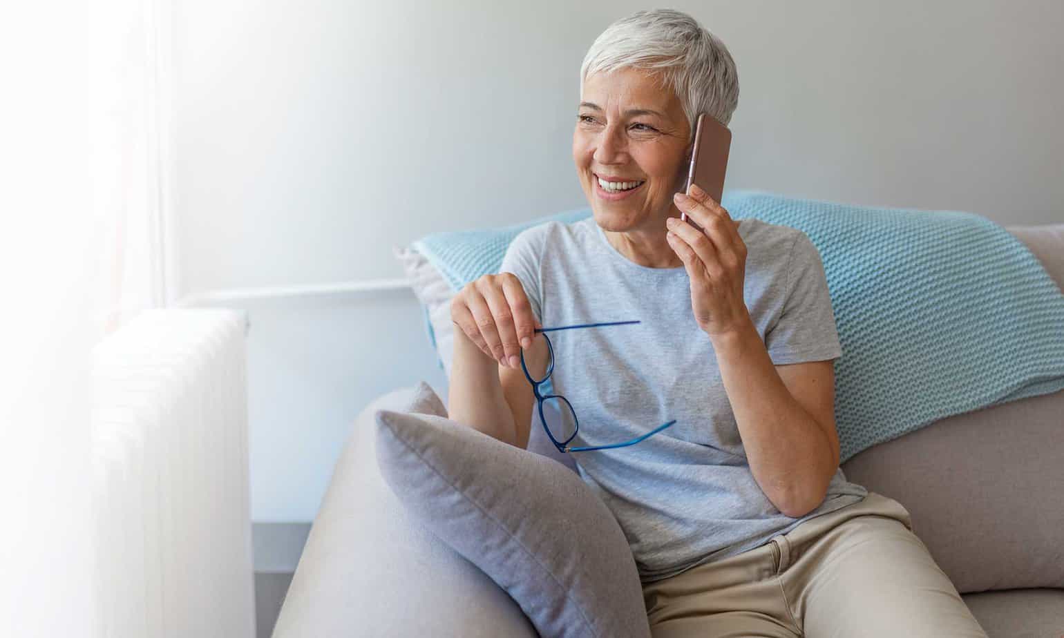 Older senior woman smiling talking on cell phone while sitting on white couch with blue blanket