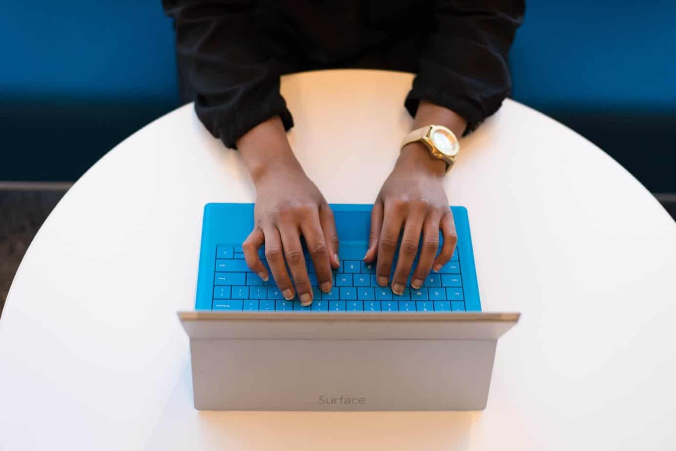 A brown-skinned woman wearing a gold watch typing on a tablet keyboard.
