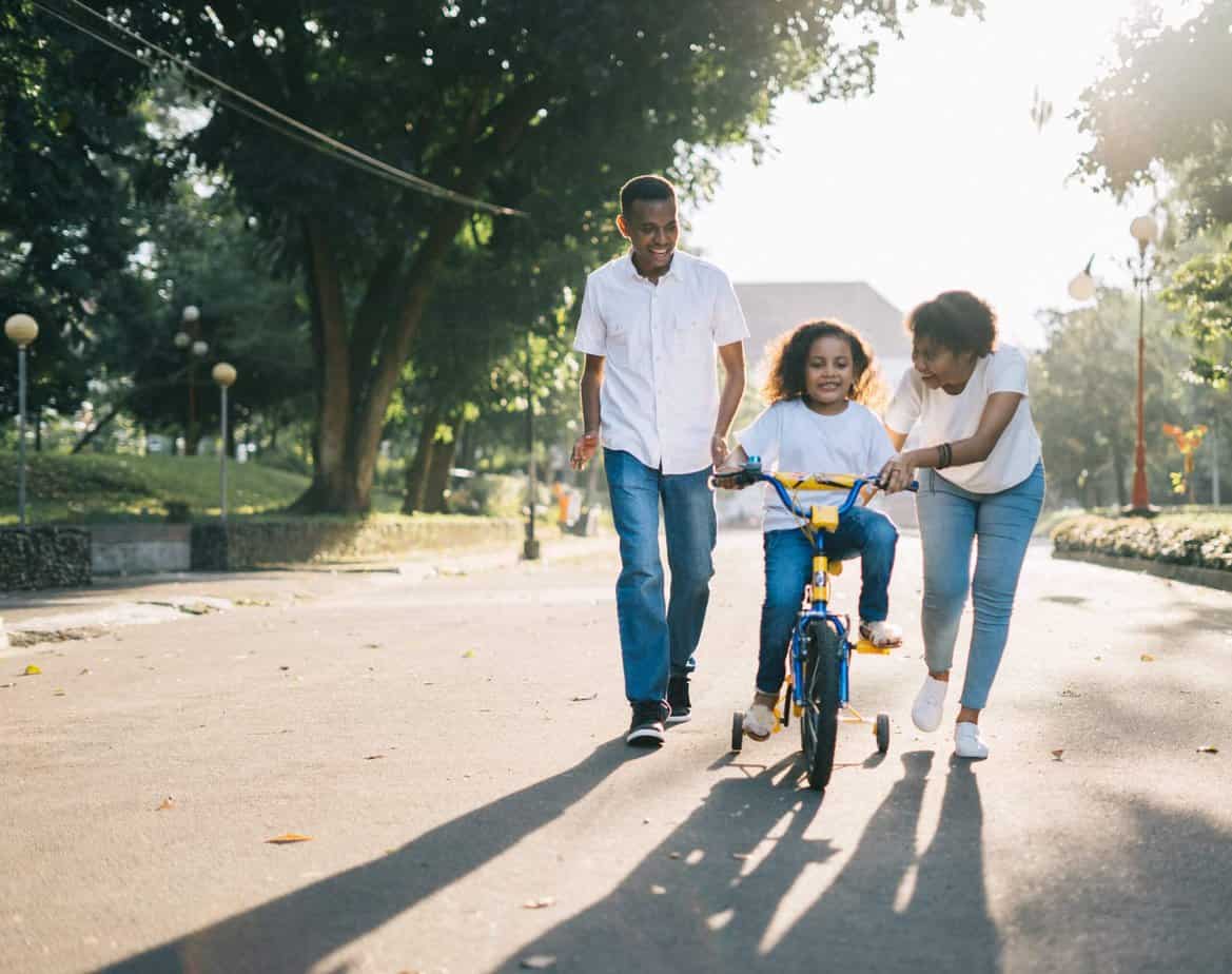 Parents teaching their daughter how to ride a bike; African-American family having fun outside.