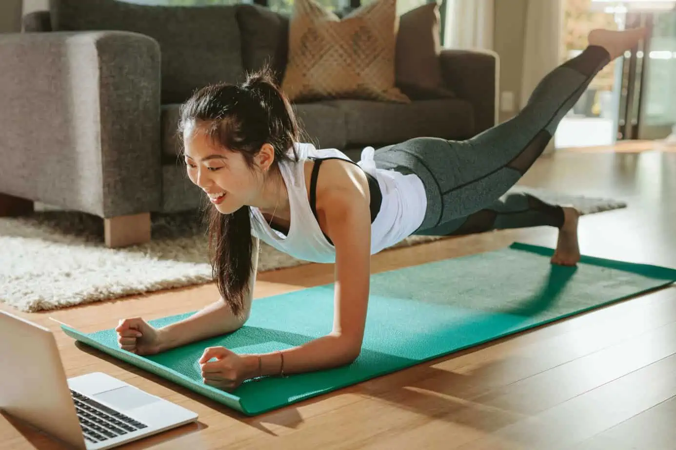 An Asian woman on a yoga mat, doing a workout with her computer in front of her.