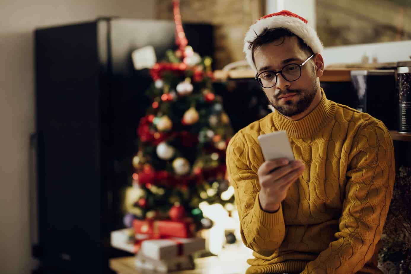 Young distraught man reading text message on cell phone while being alone at home on Christmas.