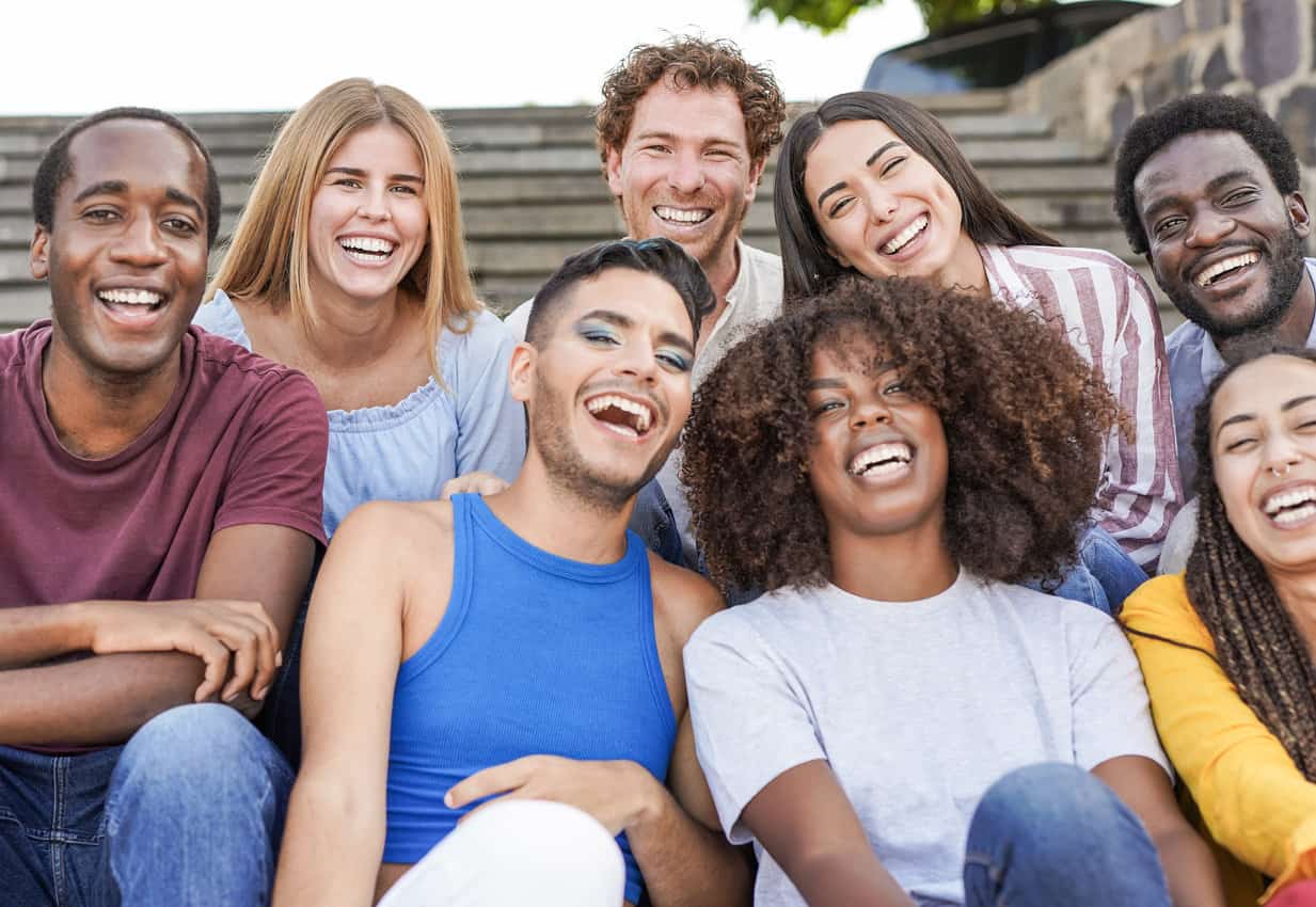 Group of mutliracial friends smiling on camera while sitting in the city - Millennial people enjoy time together outdoor - Diversity, friendship and happiness concept