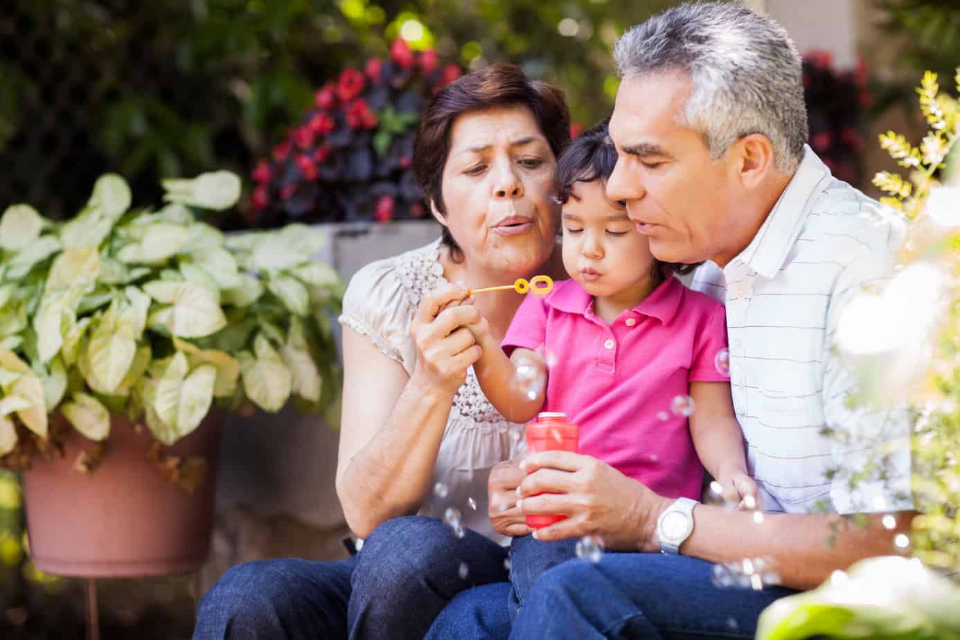 two grandparents raising grandchild sitting on bench outside blowing bubbles