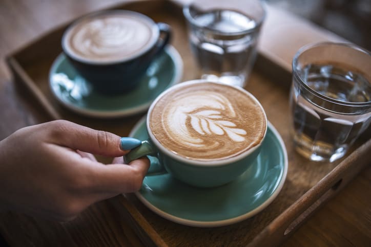 Woman's hand wearing blue nailpolish holding blue cup of latte art on wooden board in cafe